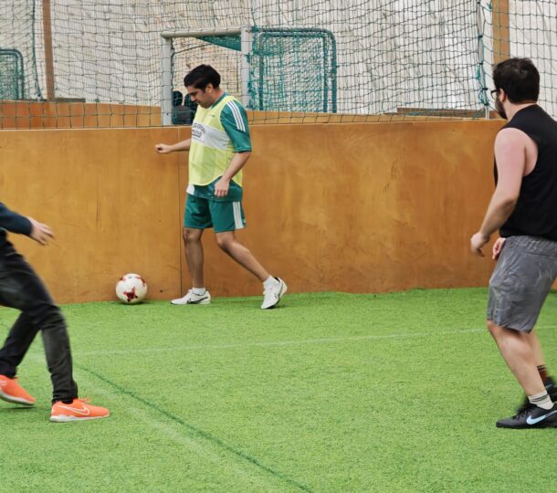 Vier Männer spielen Hallenfußball auf einem Kunstrasen; einer in Grün kontrolliert den Ball in der Nähe einer Wand, während die anderen, die in sportlicher Freizeitkleidung gekleidet sind, sich zur Verteidigung oder zum Angriff bereit machen. Ein Fußballnetz und ein Ball sind zu sehen.