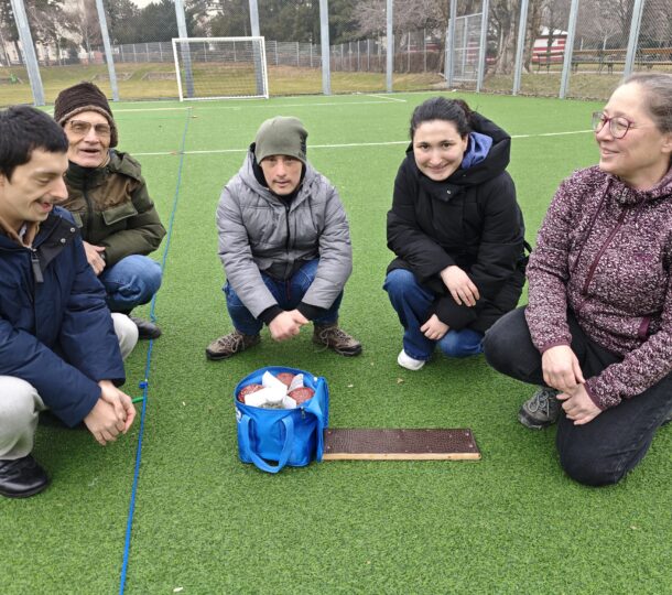 Fünf Personen in Winterkleidung knien auf einem grünen Kunstrasenplatz und haben sich um eine blaue Tasche und ein rechteckiges Holzbrett auf dem Boden versammelt. Im Hintergrund sind ein Fußballtor und ein Zaun zu sehen.