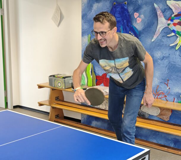 Ein Mann mit Brille und T-Shirt lächelt, während er drinnen Tischtennis spielt und neben einer bunten Wandmalerei mit Fischmotiven steht.