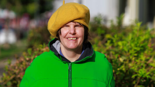 Eine lächelnde Person im Freien, die eine leuchtend gelbe Baskenmütze und eine neongrüne Jacke trägt, steht vor grünen Büschen, auf deren Gesicht die Sonne scheint.