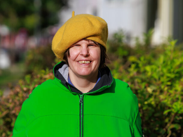 Eine lächelnde Person im Freien, die eine leuchtend gelbe Baskenmütze und eine neongrüne Jacke trägt, steht vor grünen Büschen, auf deren Gesicht die Sonne scheint.
