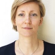 Eine Frau mit kurzen blonden Haaren und grünen Augen, die ein schwarz-weißes Oberteil und eine rechteckige Halskette mit Anhänger trägt, steht vor einer hellen Wand und schaut mit neutralem Blick direkt in die Kamera.