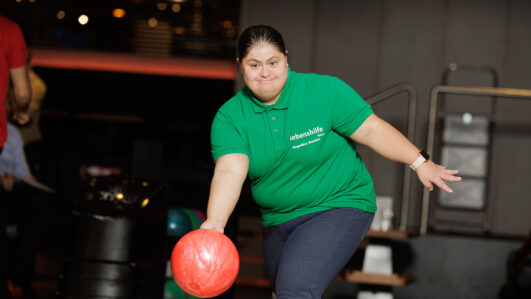 Eine Frau in einem grünen Polohemd kegelt auf einer Bowlingbahn eine rote Kugel und konzentriert sich, während sie die Kugel die Bahn hinunterwirft.