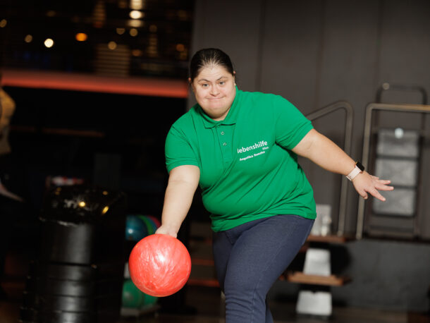 Eine Frau in einem grünen Polohemd kegelt auf einer Bowlingbahn eine rote Kugel und konzentriert sich, während sie die Kugel die Bahn hinunterwirft.
