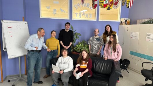 Eine Gruppe von acht Personen, von denen einige stehen und einige auf einem schwarzen Sofa sitzen, lächelt in einem farbenfrohen Büroraum mit Flipchart, Schränken und dekorativen Schildern, die von der Decke hängen.