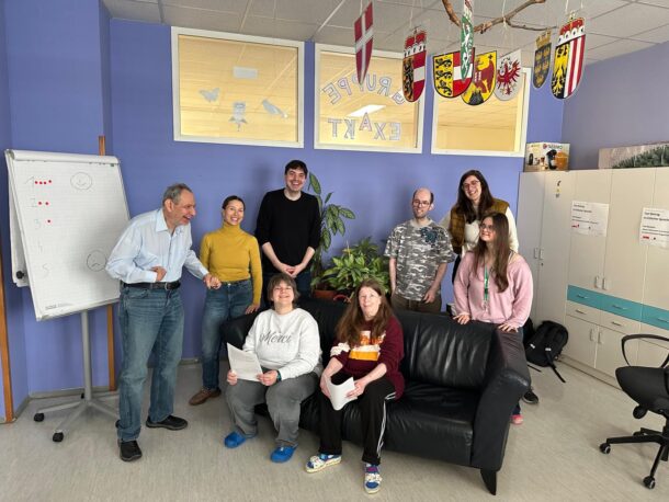 Eine Gruppe von acht Personen, von denen einige stehen und einige auf einem schwarzen Sofa sitzen, lächelt in einem farbenfrohen Büroraum mit Flipchart, Schränken und dekorativen Schildern, die von der Decke hängen.