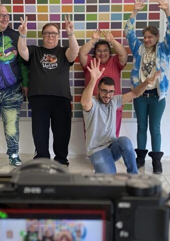 Fünf Personen posieren und machen Friedenszeichen vor einem farbenfrohen Gitterkunstwerk, während eine Kamera im Vordergrund ihr Foto in einem hellen, modernen Raum mit weißen Wänden und Böden einfängt.
