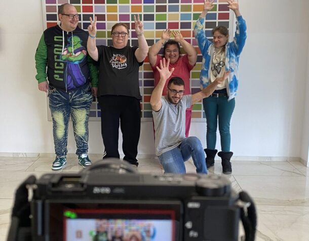 Fünf Personen posieren und machen Friedenszeichen vor einem farbenfrohen Gitterkunstwerk, während eine Kamera im Vordergrund ihr Foto in einem hellen, modernen Raum mit weißen Wänden und Böden einfängt.