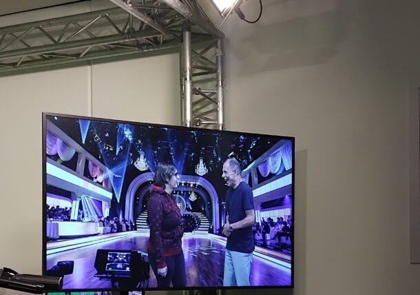 Ein Fernsehstudio mit einem großen Monitor, auf dem zwei Personen auf einer hell erleuchteten Bühne sprechen. Im Hintergrund sind Studioleuchten, Metalltraversen und Produktionsausrüstung zu sehen.