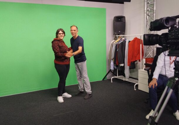 Zwei Personen stehen händchenhaltend und lächelnd vor einem Greenscreen in einem Studio. Im Raum sind eine Kamera und eine Kleiderablage zu sehen, über dem Kopf befinden sich Studioleuchten und -geräte.