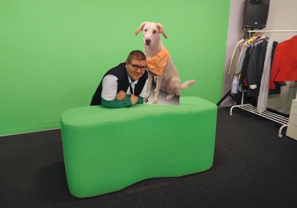 Ein lächelnder Mann lehnt an einem grünen Tisch neben einer Pappfigur eines Hundes, der ein orangefarbenes Halstuch trägt. Hinter ihnen sind ein grüner Bildschirmhintergrund und ein Kleiderständer zu sehen.