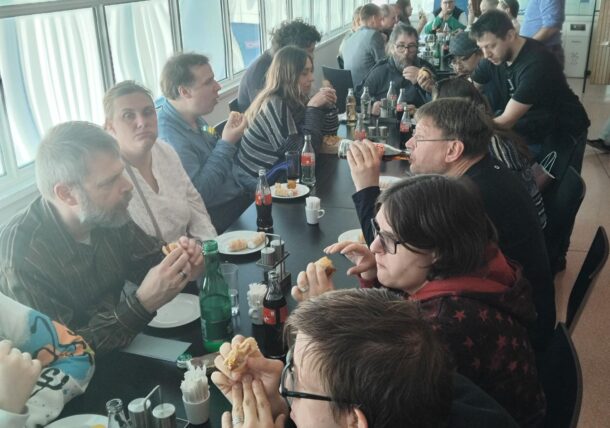Eine Gruppe von Menschen sitzt um einen langen Tisch in einer hellen, modernen Cafeteria und isst und trinkt, während sie sich unterhalten. Einige halten Sandwiches in der Hand, andere haben Limonadenflaschen oder Gläser vor sich stehen.