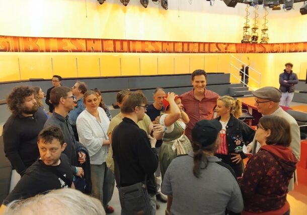 In einem hell erleuchteten Fernsehstudio mit orange-gelben Wänden und einem Banner mit der Aufschrift BISS NEUES steht eine Gruppe von Menschen zusammen und unterhält sich. Einige lächeln und unterhalten sich, während andere sich umschauen.