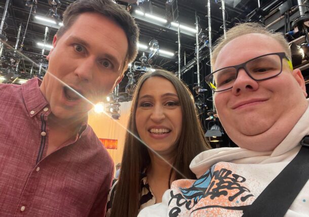 Drei Personen lächeln und posieren für ein Selfie in einem hell erleuchteten Studio, in dem Lichter und Kabel über dem Kopf zu sehen sind. Die Person links trägt ein rotes Hemd, die Person in der Mitte hat lange dunkle Haare und die Person rechts trägt eine Brille und einen weißen Kapuzenpullover.