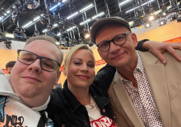 Drei Personen, zwei Männer und eine Frau, lächeln, während sie für ein Selfie in einem hell erleuchteten Studio posieren, in dem Deckenleuchten und Geräte im Hintergrund zu sehen sind. Ein Mann trägt eine Brille und eine Mütze, der andere eine Brille und einen Kapuzenpulli.