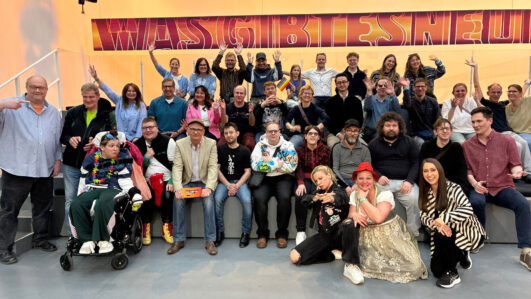 Eine große Gruppe von Menschen unterschiedlichen Alters und unterschiedlicher Stile, einige sitzend, andere stehend, posiert gemeinsam auf einer Tribüne, lächelt und macht verschiedene feierliche Gesten vor einer farbenfrohen Kulisse.