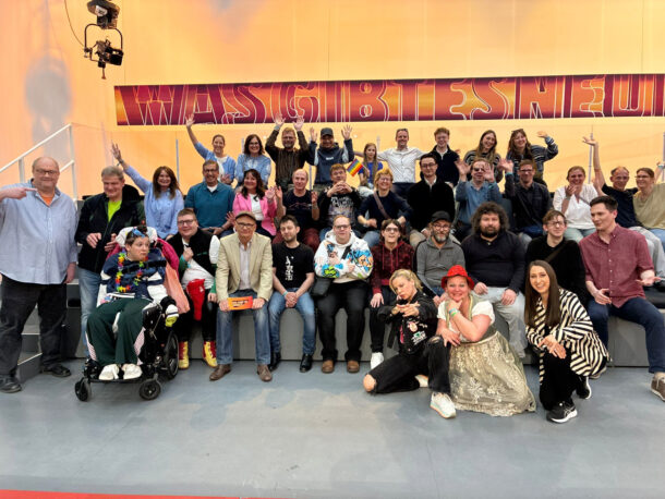 Eine große Gruppe von Menschen unterschiedlichen Alters und unterschiedlicher Stile, einige sitzend, andere stehend, posiert gemeinsam auf einer Tribüne, lächelt und macht verschiedene feierliche Gesten vor einer farbenfrohen Kulisse.