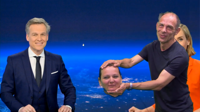 Ein Mann im Anzug sitzt an einem Schreibtisch und lächelt, während ein anderer Mann in einem schwarzen Hemd eine große, realistische Maske eines Frauenkopfes in der Hand hält - Teil einer Ausflug ORF-Veranstaltung. Eine Frau in Orange ist teilweise vor einem Nachthimmel über der Erde zu sehen.