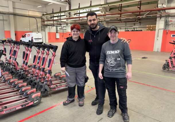 Drei Personen stehen lächelnd in einer Lagerhalle, umgeben von Reihen pinker Elektroroller. Der Hintergrund besteht aus roten Wänden, einem weißen Lieferwagen und Industriebeleuchtung über dem Kopf.