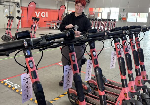Eine Person mit kurzen roten Haaren steht in einer Lagerhalle inmitten von Reihen pinkfarbener Elektroroller und befestigt Schilder an den Lenkstangen. Die Umgebung ist industriell, mit Geräten und Bannern im Hintergrund.