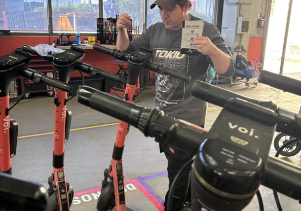 Eine Person mit Mütze steht zwischen mehreren roten Voi-Elektrorollern in einer Werkstatt, hält ein Stück Papier in der Hand und scheint die Roller zu inspizieren oder an ihnen zu arbeiten. Im Hintergrund sind Werkzeuge und Geräte zu sehen.