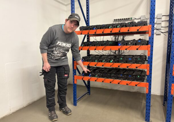 Eine Person, die eine Mütze und dunkle Kleidung trägt, steht in einem Raum mit Betonboden neben Metallregalen, die mit elektronischen Geräten gefüllt sind, bei denen es sich möglicherweise um Geräte zum Schürfen von Kryptowährungen handelt.