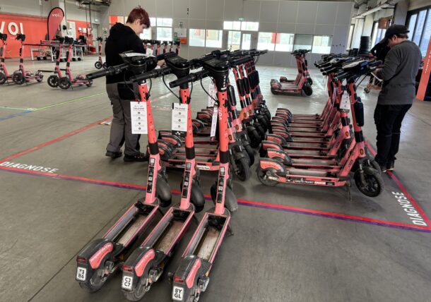 Mehrere Reihen roter Elektroroller sind in Innenräumen innerhalb markierter Zonen mit der Aufschrift DIAGNOSE geparkt, während zwei Personen einige der Roller inspizieren oder an ihnen arbeiten. Die Umgebung scheint eine Wartungs- oder Lagereinrichtung zu sein.