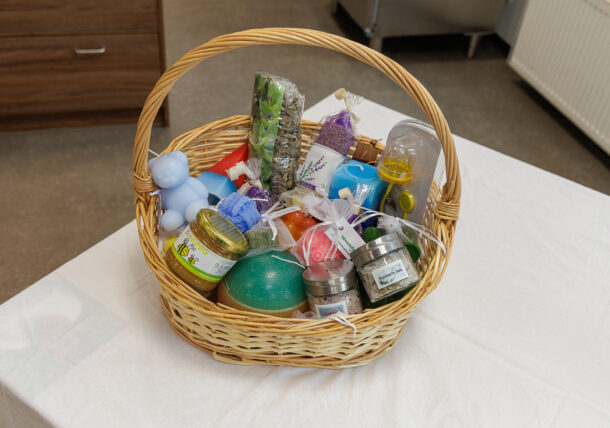 Ein Geschenkkorb aus Weidengeflecht, gefüllt mit verschiedenen farbenfrohen Gegenständen, darunter Gläser, Seifen, ein Objekt in Form eines blauen Bären, Badebomben und kleine Tüten mit Badesalz, ausgestellt auf einer weißen Tischdecke.
