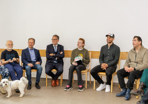Eine Gruppe von sechs Personen sitzt in einem Halbkreis auf Holzstühlen, lächelt und unterhält sich. Ein kleiner weißer Hund steht vor ihnen auf dem Boden. Der Hintergrund ist eine schlichte weiße Wand.
