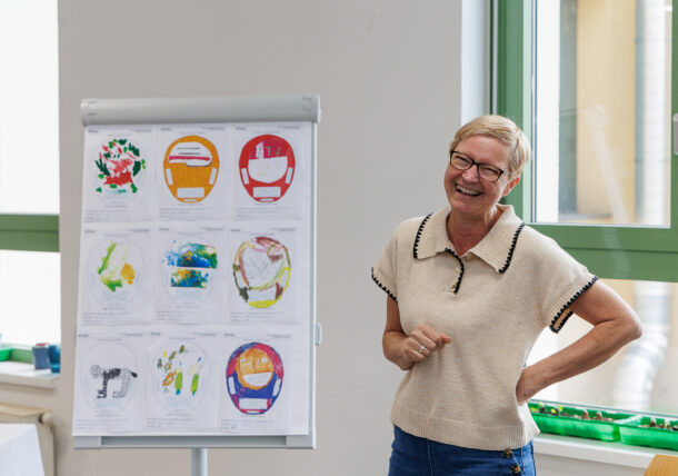 Eine Frau mit Brille und einem hellen kurzärmeligen Pullover steht lächelnd neben einem Flipchart mit bunten Kreiszeichnungen in einem hellen Raum mit großen Fenstern.