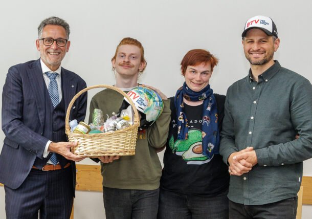 Vier Personen stehen lächelnd zusammen. Die Person links im Anzug hält einen großen Geschenkkorb mit Getränken und Snacks in der Hand. Die anderen sind leger gekleidet, einer trägt eine weiße Mütze mit der Aufschrift 