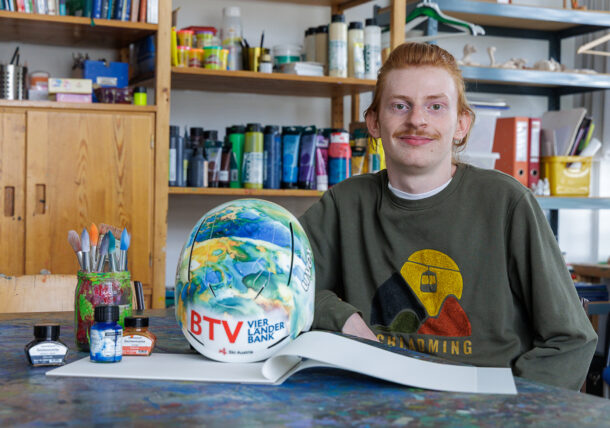 Eine lächelnde Person mit rötlichem Haar und Schnurrbart sitzt an einem farbenfrohen Schreibtisch mit Kunstzubehör, einem bemalten Helm und einem aufgeschlagenen Skizzenbuch in einem mit Farben, Pinseln und Regalen gefüllten Kunstatelier im Hintergrund.