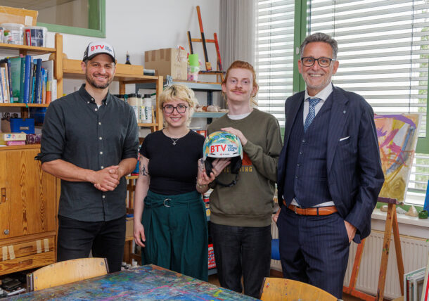 Vier Personen stehen in einem Haus und lächeln in die Kamera. Eine Person hält einen bunten Helm mit der Aufschrift BTV. Im Hintergrund sind Bücherregale, Kunstgegenstände und große Fenster mit Jalousien zu sehen.