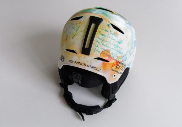 Ein weißer Skihelm mit bunten abstrakten Motiven und handgeschriebenen Botschaften in grüner und schwarzer Tinte auf der Rückseite. Der Helm trägt den Namen JOHANNES STROLZ und hat einen verstellbaren Kinnriemen.