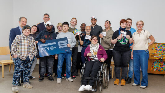 Eine Gruppe lächelnder Menschen, darunter auch Menschen mit Behinderungen, posieren gemeinsam in einem Raum. Einige halten Karten und ein großes blaues BTV-Schild hoch. Eine Person sitzt in einem Rollstuhl. Alle sehen glücklich und engagiert aus.