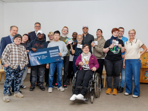 Eine Gruppe lächelnder Menschen, darunter auch Menschen mit Behinderungen, posieren gemeinsam in einem Raum. Einige halten Karten und ein großes blaues BTV-Schild hoch. Eine Person sitzt in einem Rollstuhl. Alle sehen glücklich und engagiert aus.