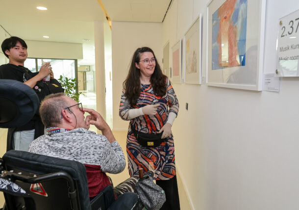Eine Frau in einem gemusterten Kleid gestikuliert in Richtung eines gerahmten Kunstwerks an der Wand und spricht einen Mann im Rollstuhl und einen anderen Mann an, die hinter ihr stehen und beide in einem hellen Galerieraum beobachten und fotografieren.