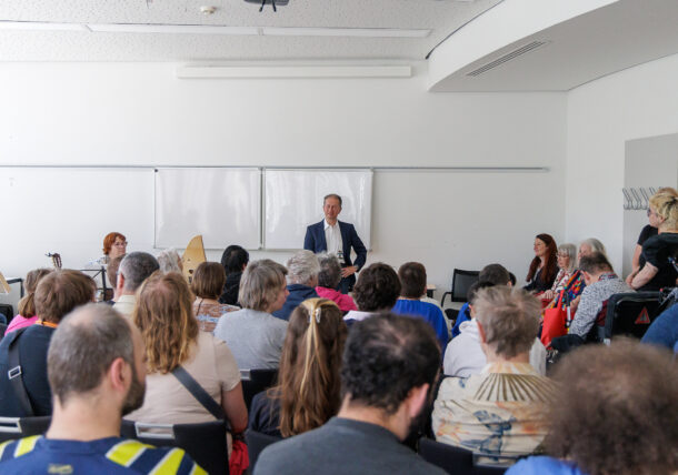 Ein Mann im Anzug steht vorne in einem Klassenzimmer und spricht zu einem sitzenden Publikum. Mehrere Personen sitzen neben ihm, und Musikinstrumente sind zu sehen. Der Raum ist hell, mit weißen Wänden und einem Whiteboard.