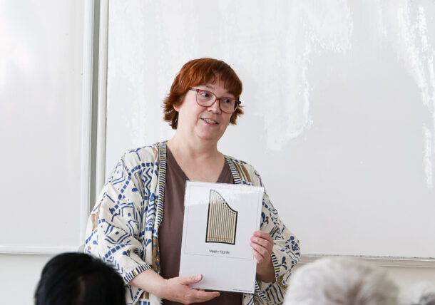 Eine Frau mit kurzen roten Haaren und Brille steht vor einem Whiteboard und hält eine Mappe mit einem Bild von Panflöten und der Aufschrift Valtin-Panflöte hoch.