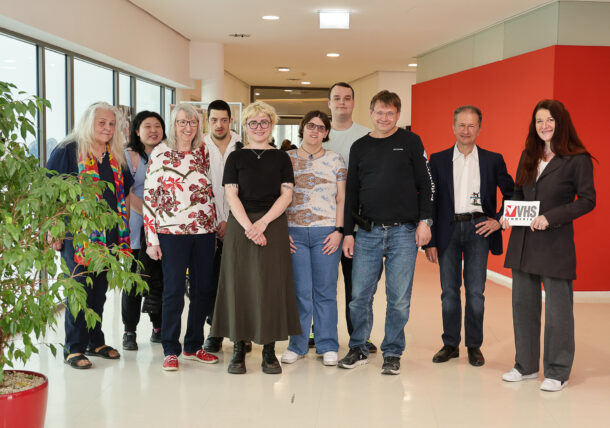 Eine Gruppe von zehn Personen steht in einem Innenraum zusammen und lächelt in die Kamera. Eine Person auf der rechten Seite hält ein Schild mit der Aufschrift VHS. Im Hintergrund sind eine große rote Wand und Fenster zu sehen, und im Vordergrund steht eine grüne Pflanze.
