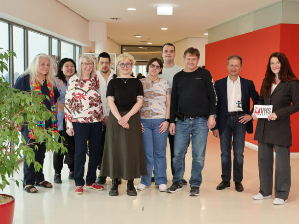 Eine Gruppe von zehn Personen steht in einem Innenraum zusammen und lächelt in die Kamera. Eine Person auf der rechten Seite hält ein Schild mit der Aufschrift VHS. Auf der linken Seite steht eine grüne Pflanze, und im Hintergrund sind große Fenster und eine rote Akzentwand zu sehen.