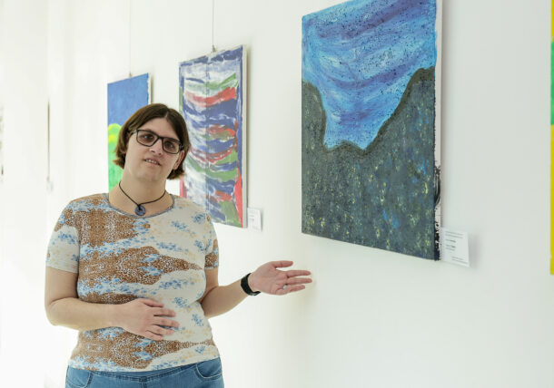 Eine Person mit kurzen braunen Haaren und Brille steht in einer Galerie und deutet mit einer Geste auf ein abstraktes blau-graues Gemälde an der Wand, das den Betrachter einlädt, Kunst mit allen Sinnen zwischen den anderen bunten Kunstwerken, die daneben ausgestellt sind, zu erleben.