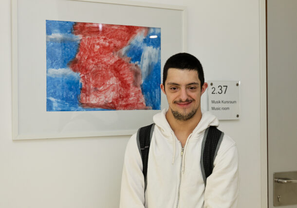 Ein junger Mann mit Down-Syndrom, der einen weißen Kapuzenpulli und einen Rucksack trägt, steht lächelnd vor einem farbenfrohen abstrakten Kunstwerk und einem Schild mit der Aufschrift 2.37 Music room an einer weißen Wand.