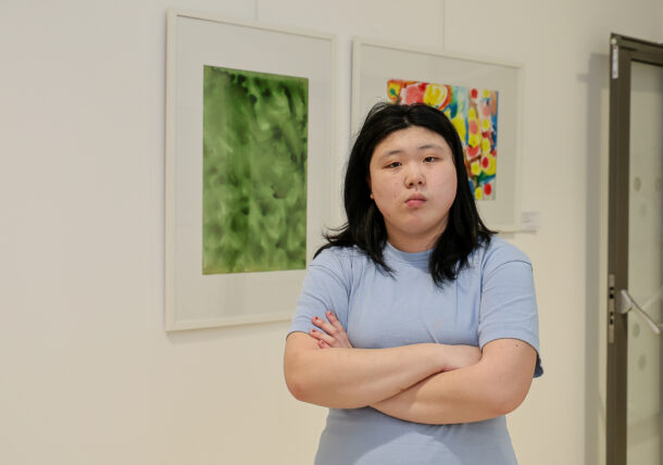 Eine Person mit langen dunklen Haaren und einem hellblauen Hemd steht mit verschränkten Armen vor zwei farbenfrohen abstrakten Gemälden an einer weißen Wand in einer Galerie.