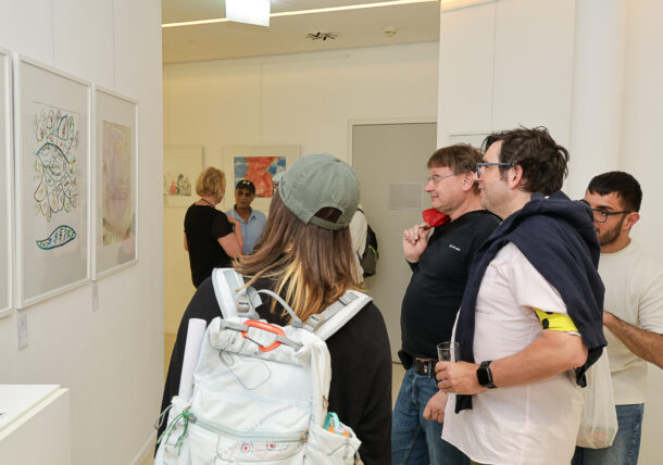 Eine Gruppe von Personen betrachtet Kunstwerke, die in einer hell erleuchteten Galerie an weißen Wänden ausgestellt sind. Einige sehen sich die Kunstwerke genau an, während andere sich unterhalten. Eine Person hat einen weißen Rucksack, eine andere hält ein Getränk in der Hand.