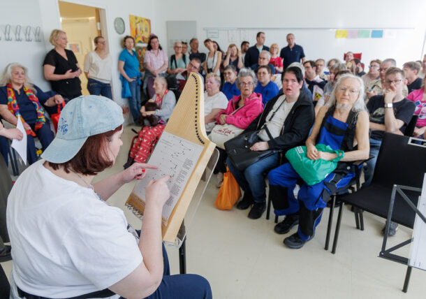 Eine Frau mit umgekehrter Mütze spielt ein Saiteninstrument, während sie Noten liest, und tritt vor einem sitzenden oder stehenden Publikum in einem hellen Klassenzimmer oder Gemeindesaal auf.