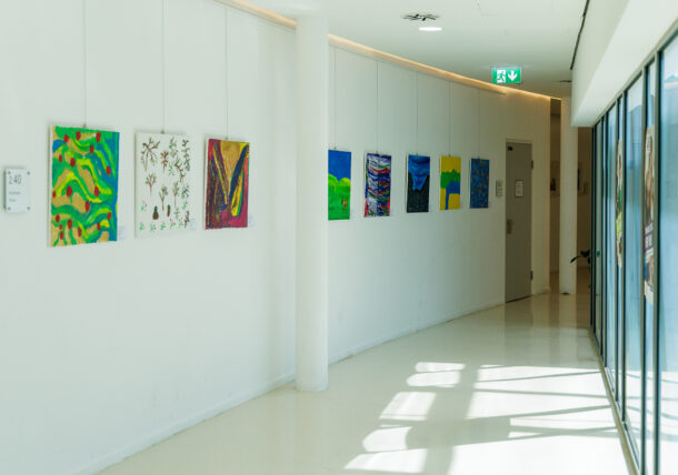 Ein heller Flur mit bunten abstrakten Gemälden an einer weißen Wand. Das Sonnenlicht fällt durch große Fenster auf der rechten Seite ein und wirft Schatten auf den glänzenden Boden. Ein Notausgangsschild ist über einer Tür am Ende des Flurs zu sehen.
