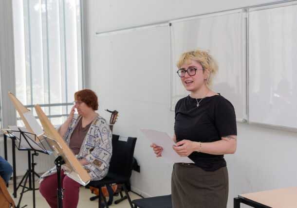 Eine Person mit kurzen blonden Haaren und Brille steht in einem Klassenzimmer, hält Papiere in der Hand und spricht. Eine andere Person sitzt in der Nähe und hält sich die Hand vor das Gesicht. Im Hintergrund sind Notenständer und eine Gitarre zu sehen.