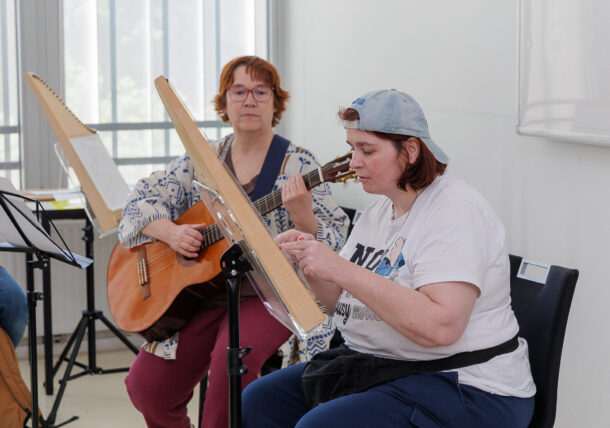 Zwei Frauen sitzen in einem Haus und spielen Streichinstrumente, eine auf der Gitarre, die andere auf einem harfenähnlichen Instrument. Sie konzentrieren sich auf ihre Musik, während vor ihnen Notenständer stehen und das Licht durch hohe Fenster strömt.