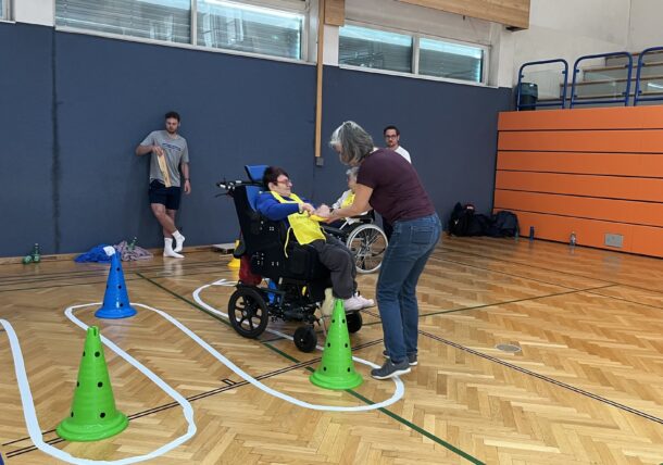 Eine Frau hilft einer Person im Rollstuhl bei der Bewältigung eines Hindernisparcours mit farbigen Kegeln in einer Sporthalle. Mehrere Personen stehen in der Nähe und beobachten das Geschehen.